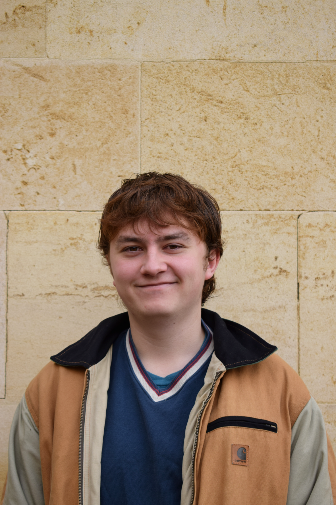 A young white, cis-man looking face on at the camera, smiling. Jamie has tousled brown hair, and is wearing a beige and green jacket over a blue v-neck sweater. He is standing in front of a sandstone wall.