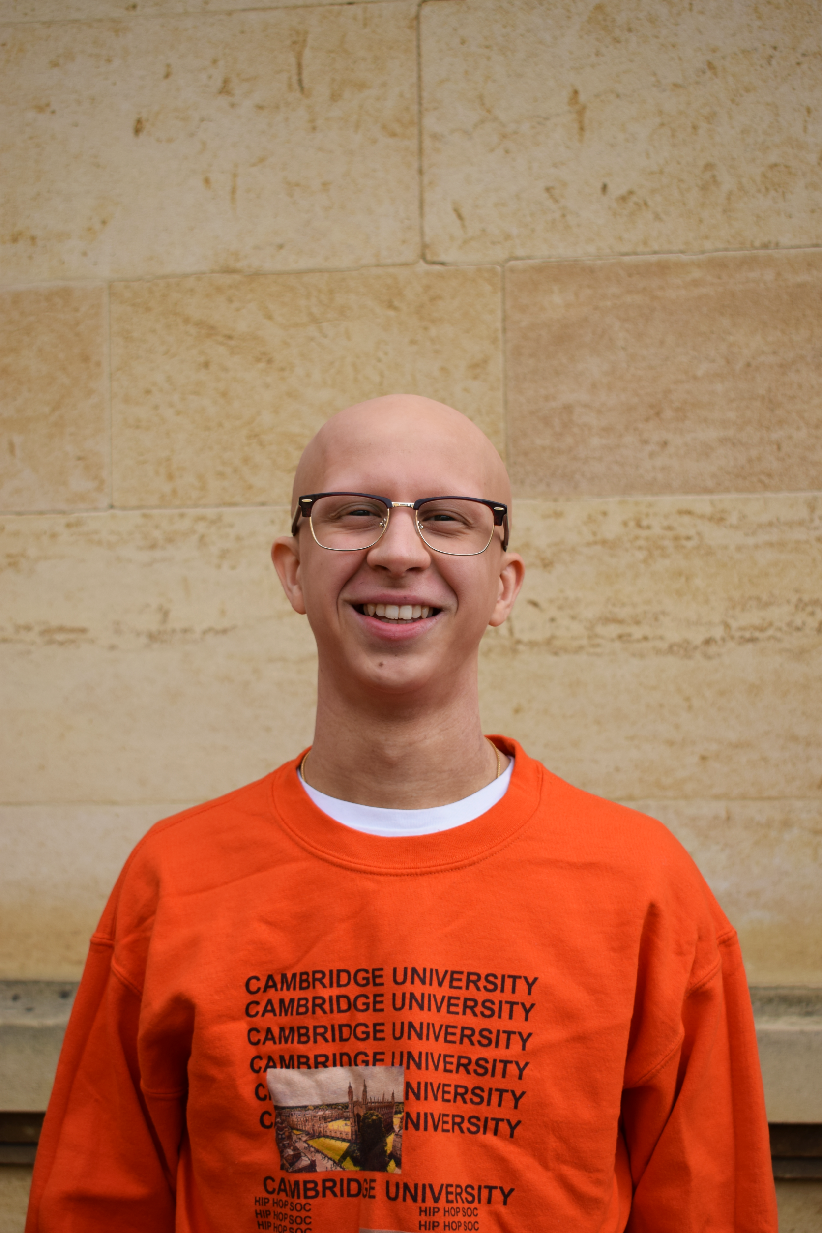 A young, bald white man in an orange jumper, standing in front of a stone wall