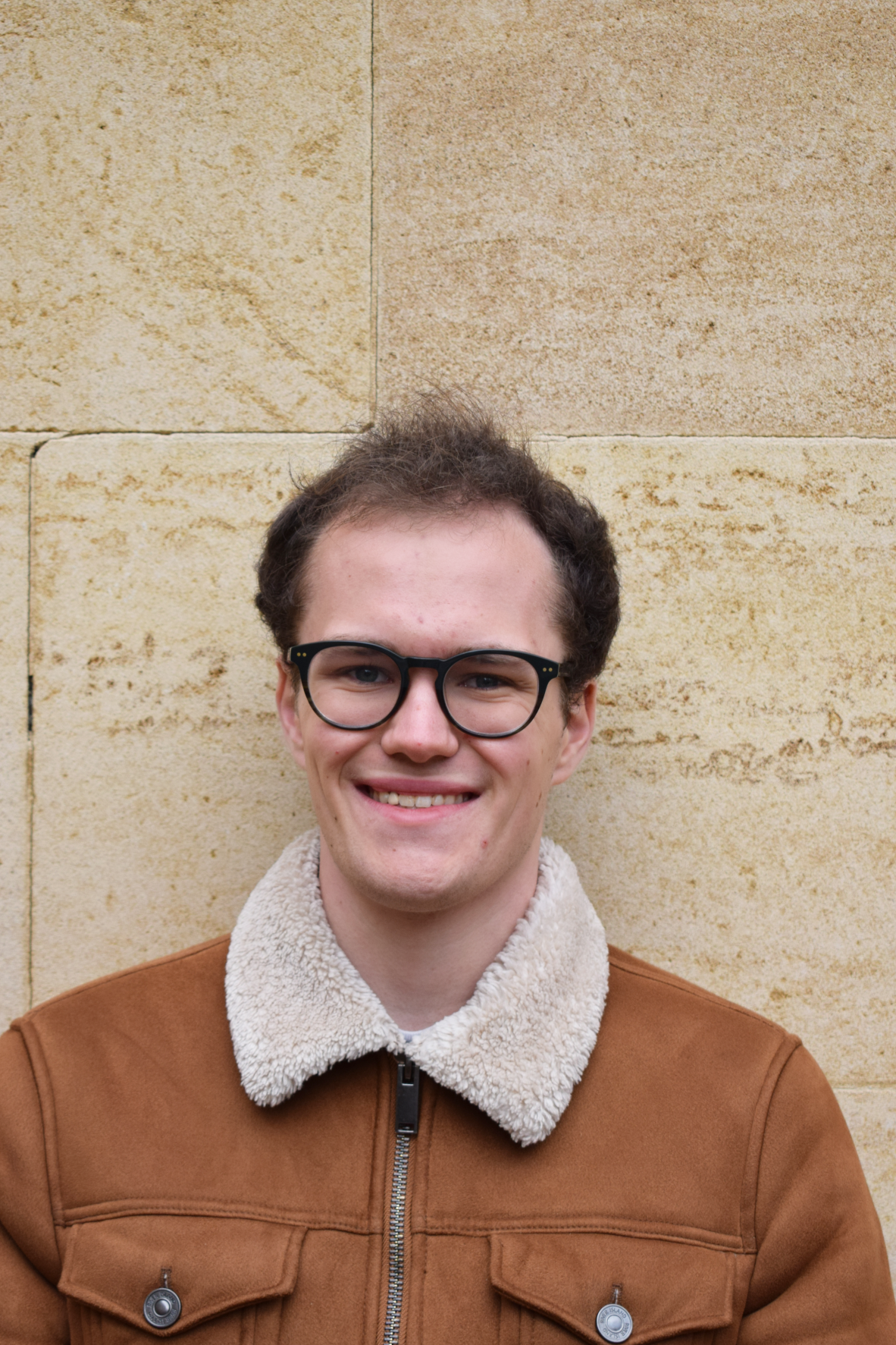 Will, a young cis-gender white man, facing head-on and smiling at the camera. He has short, dark brown hair, glasses, and is wearing a light brown sheepskin coat.