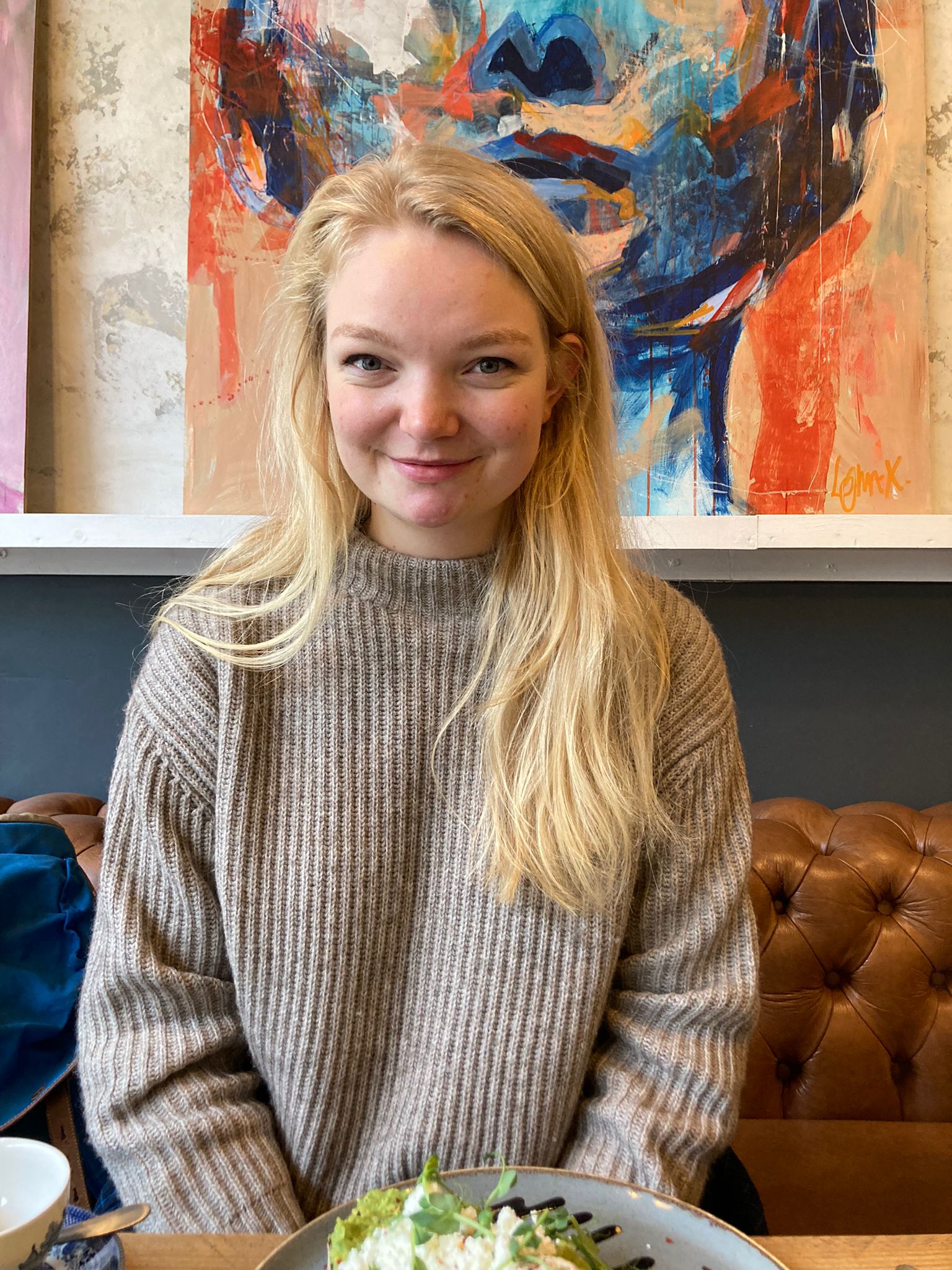 Georgia is smiling head-on at the camera. There is a sandstone wall behind her, with colourful curtains. She is a white cis-gender woman, has chest-length blonde hair, and is wearing a grey sweater.