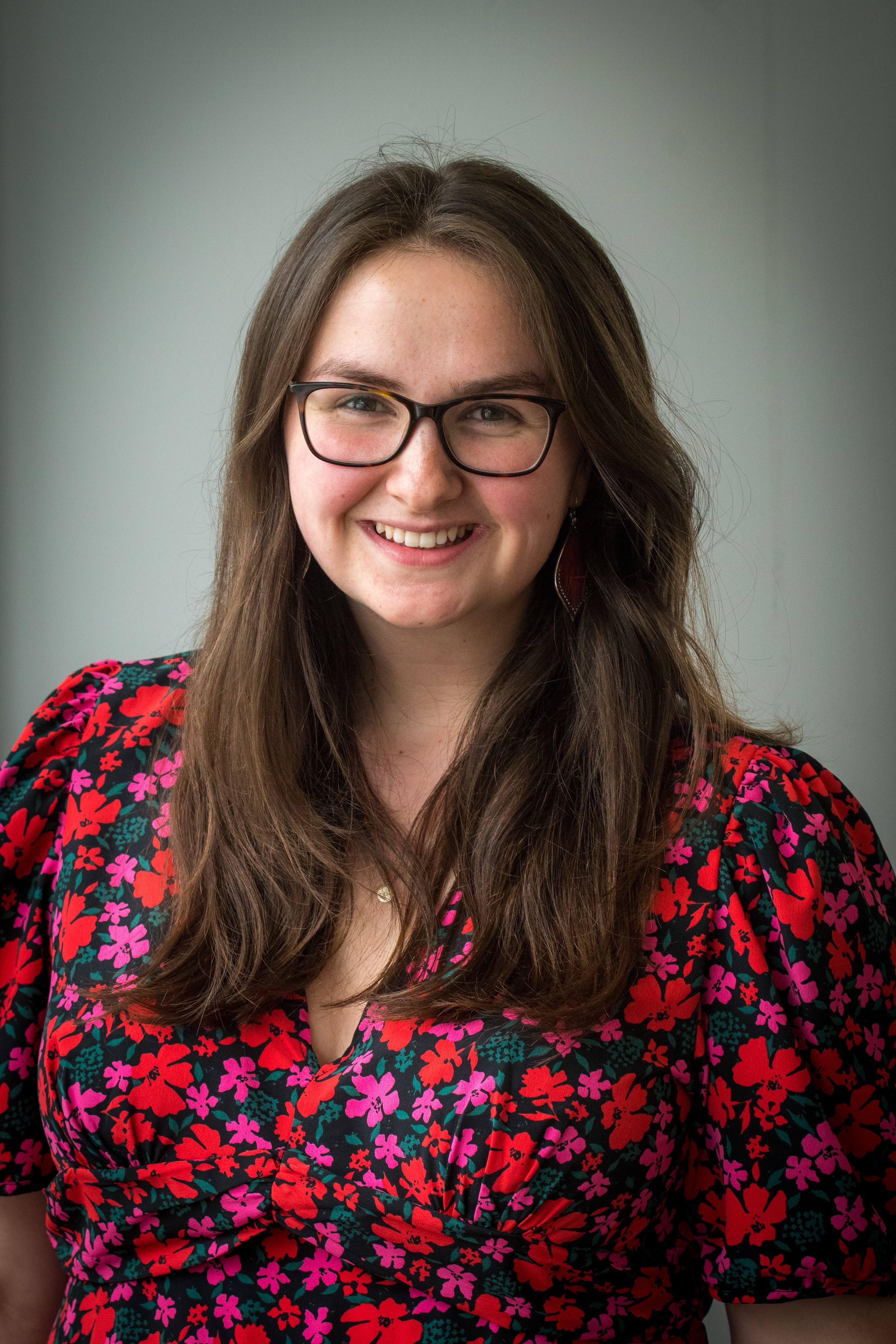 Kirstie is smiling head-on at the camera. There is a light grey background for her headshot. She is a white cis-gender woman, has chest-length brown hair, rectangular glasses, a round face, brown eyes, long red earrings, and is wearing a multi-coloured floral dress.